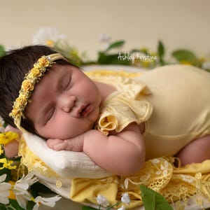 May include: A newborn baby wearing a yellow headband with floral accents and a matching yellow onesie, resting on a white pillow surrounded by flowers. The baby is nestled in a bed of white and yellow flowers.