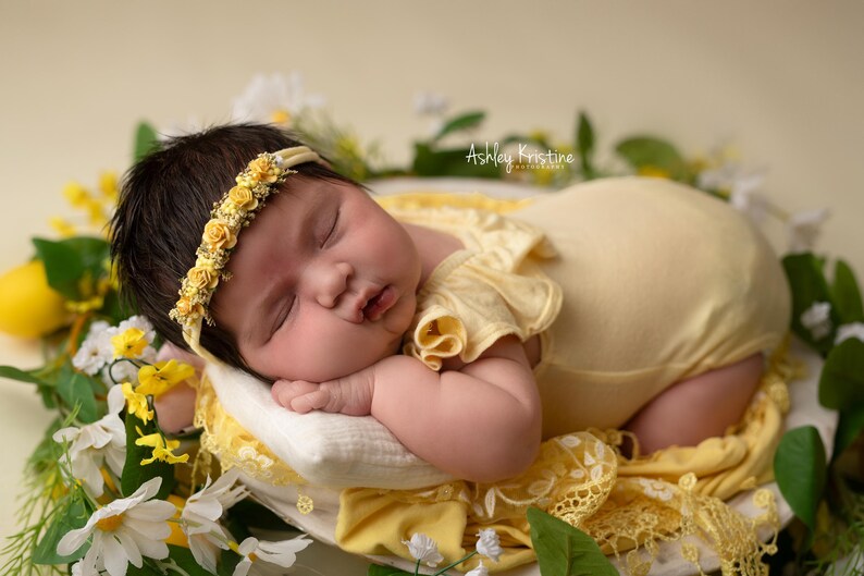 May include: A newborn baby wearing a yellow headband with floral accents and a matching yellow onesie, resting on a white pillow surrounded by flowers. The baby is nestled in a bed of white and yellow flowers.