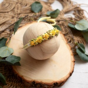 May include: A yellow floral headband with small yellow roses and greenery is displayed on a wooden sphere. The headband is set on a wooden slice, with eucalyptus leaves and a woven background.