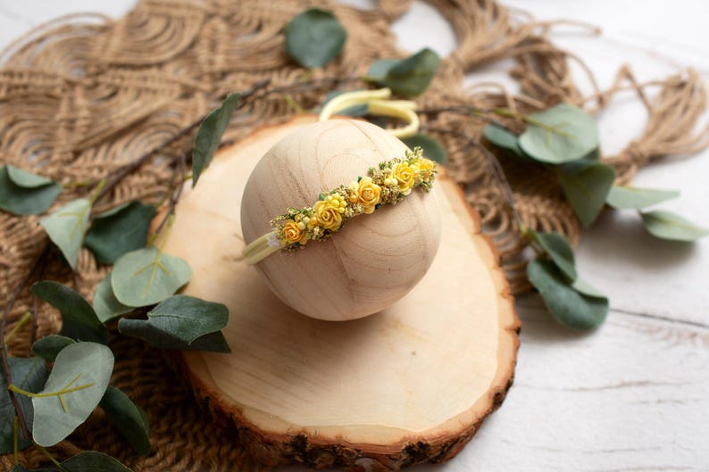 May include: A yellow floral headband with small yellow roses and greenery is displayed on a wooden sphere. The headband is set on a wooden slice, with eucalyptus leaves and a woven background.