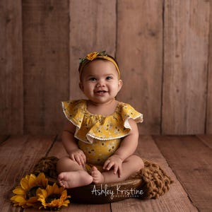 Op de afbeelding: Een lachende baby in een gele bloemenromper en bijpassende hoofdband. De baby zit op een bruine gebreide deken met zonnebloemen en een houten achtergrond. De romper heeft een witte kanten rand.