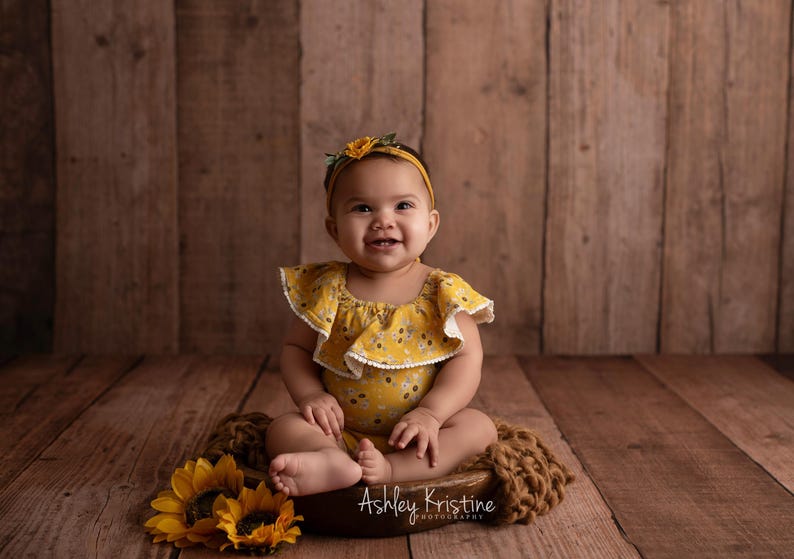 Op de afbeelding: Een lachende baby in een gele bloemenromper en bijpassende hoofdband. De baby zit op een bruine gebreide deken met zonnebloemen en een houten achtergrond. De romper heeft een witte kanten rand.