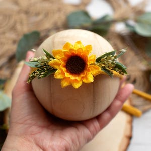 Op de afbeelding: Een houten bol versierd met een felgele zonnebloem en groen. De zonnebloem heeft een donkerbruin hart en felgele bloemblaadjes. De bloemstuk is waarschijnlijk een haarband of haaraccessoire.
