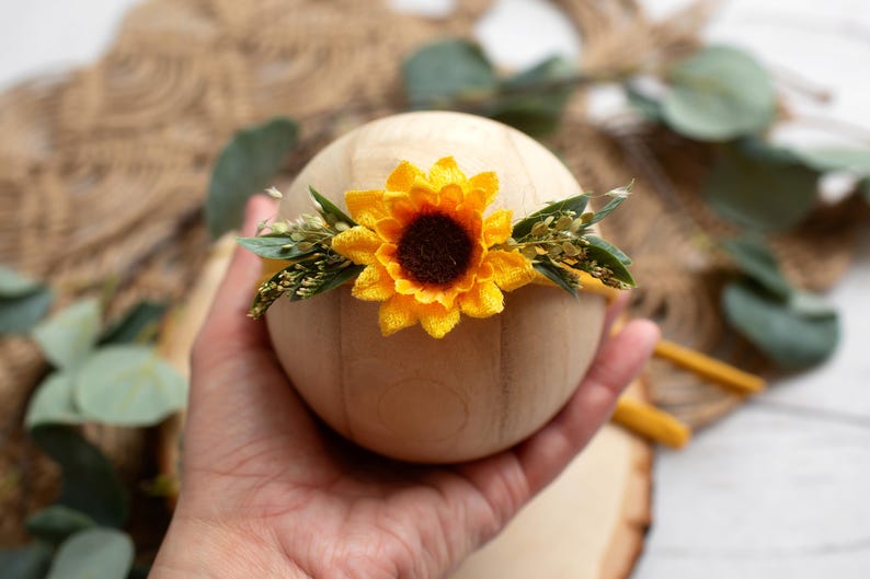 Op de afbeelding: Een houten bol versierd met een felgele zonnebloem en groen. De zonnebloem heeft een donkerbruin hart en felgele bloemblaadjes. De bloemstuk is waarschijnlijk een haarband of haaraccessoire.