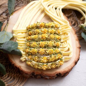 May include: A collection of yellow floral headbands arranged on a wooden slice, surrounded by strands of yellow material. The headbands are adorned with small yellow roses and greenery, creating a delicate and natural aesthetic.