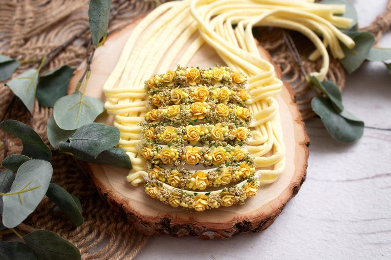 May include: A collection of yellow floral headbands arranged on a wooden slice, surrounded by strands of yellow material. The headbands are adorned with small yellow roses and greenery, creating a delicate and natural aesthetic.