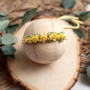 May include: A baby headband featuring small yellow roses and greenery. The floral arrangement is set on a light-colored band. The headband is displayed on a wooden sphere, with a wooden slice and greenery in the background.