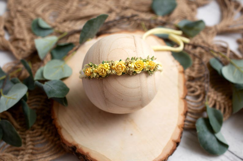 May include: A baby headband featuring small yellow roses and greenery. The floral arrangement is set on a light-colored band. The headband is displayed on a wooden sphere, with a wooden slice and greenery in the background.