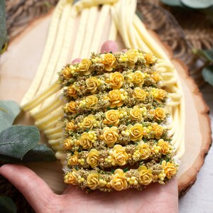 May include: A floral headband featuring rows of small yellow roses and tiny yellow buds. The headband is held in a hand, with a soft yellow fabric and wooden background. The flowers are a vibrant yellow.