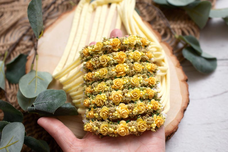 May include: A floral headband featuring rows of small yellow roses and tiny yellow buds. The headband is held in a hand, with a soft yellow fabric and wooden background. The flowers are a vibrant yellow.