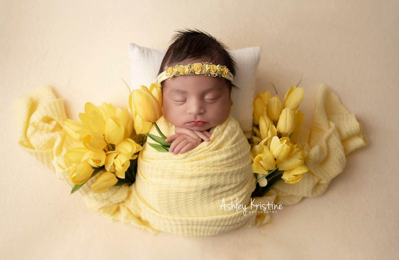 May include: A newborn baby swaddled in a yellow blanket, resting on a white pillow, and wearing a yellow floral headband. The baby is surrounded by yellow tulips and a yellow blanket. The image is signed "Ashley Kristine Photography."