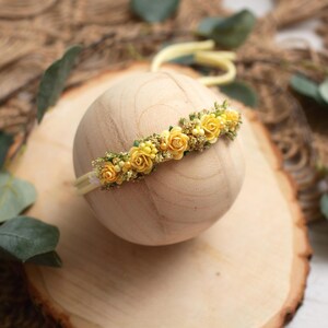 May include: A yellow floral headband with small rose accents. The headband is set on a wooden sphere, with a light yellow elastic band. The background includes greenery and a wooden surface.