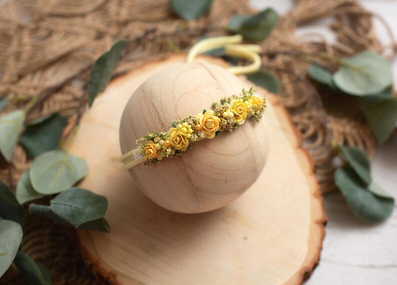 May include: A yellow floral headband with small rose accents. The headband is set on a wooden sphere, with a light yellow elastic band. The background includes greenery and a wooden surface.