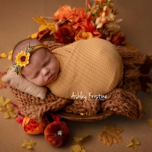 Op de afbeelding: Een slapende pasgeboren baby, gewikkeld in een gele deken, met een zonnebloemhoofdband, rustend op een geweven bruin oppervlak. De baby is omringd door herfstdecoraties, waaronder bloemen en bladeren, wat een warme, herfstachtige sc&egrave;ne cre&euml;ert.