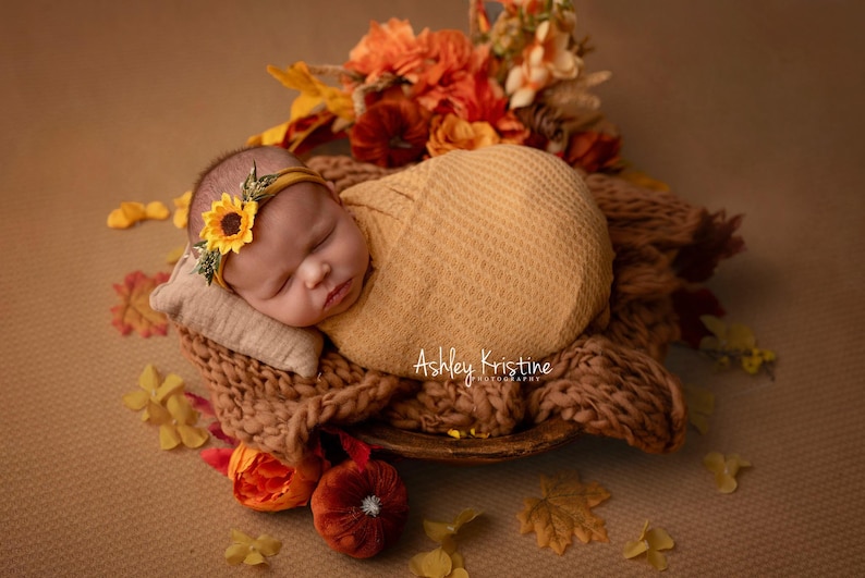 Op de afbeelding: Een slapende pasgeboren baby, gewikkeld in een gele deken, met een zonnebloemhoofdband, rustend op een geweven bruin oppervlak. De baby is omringd door herfstdecoraties, waaronder bloemen en bladeren, wat een warme, herfstachtige sc&egrave;ne cre&euml;ert.