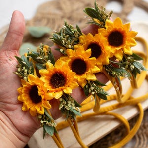 Op de afbeelding: Een verzameling gele zonnebloem haarbanden met donkerbruine centra en groene bladeren. De haarbanden worden in een hand gehouden, waarbij de heldere kleuren en het bloemendesign worden benadrukt. Een perfect accessoire voor baby's en peuters.