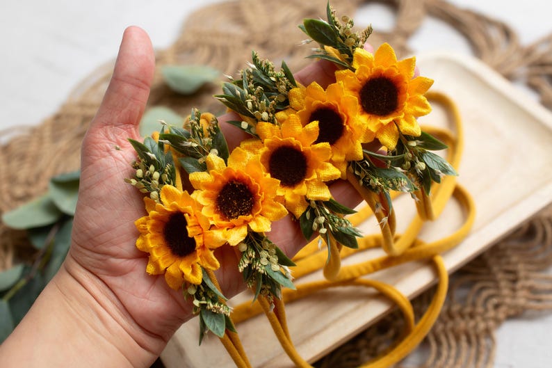 Op de afbeelding: Een verzameling gele zonnebloem haarbanden met donkerbruine centra en groene bladeren. De haarbanden worden in een hand gehouden, waarbij de heldere kleuren en het bloemendesign worden benadrukt. Een perfect accessoire voor baby's en peuters.