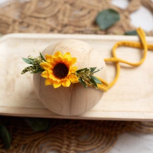 Op de afbeelding: Een houten bol versierd met een heldergele zonnebloem en groen, rustend op een licht houten dienblad. Een mosterdkleurige hoofdband is om de bol geslingerd. De opstelling staat tegen een neutrale, getextureerde achtergrond.