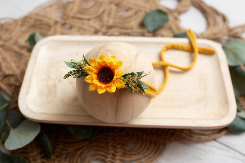 Op de afbeelding: Een houten bol versierd met een heldergele zonnebloem en groen, rustend op een licht houten dienblad. Een mosterdkleurige hoofdband is om de bol geslingerd. De opstelling staat tegen een neutrale, getextureerde achtergrond.