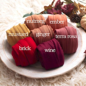 Op de afbeelding: Een verzameling stoffen rollen in verschillende herfstkleuren, waaronder mosterd, ember en wijn, gerangschikt op een witte plaat. De afbeelding is versierd met herfstbladeren en een kleine pompoen, wat een seizoensgebonden thema creëert.