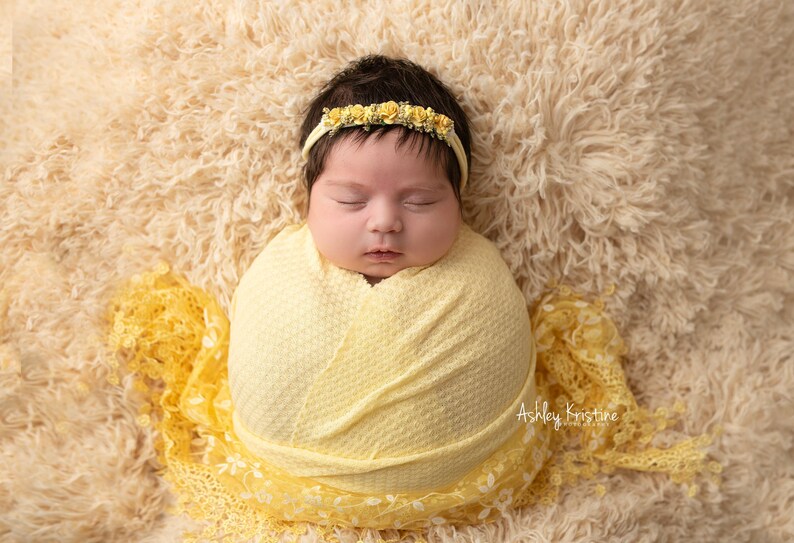 May include: A newborn baby wrapped in a yellow blanket and wearing a yellow floral headband. The baby is lying on a fluffy, cream-colored blanket. The image is a close-up shot of the baby, with the focus on the baby's face.