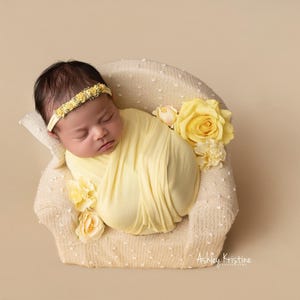 May include: A newborn baby wrapped in a soft yellow blanket, resting in a small, cream-colored chair. The baby wears a yellow floral headband. Yellow roses and other flowers decorate the chair. The background is a solid beige color.
