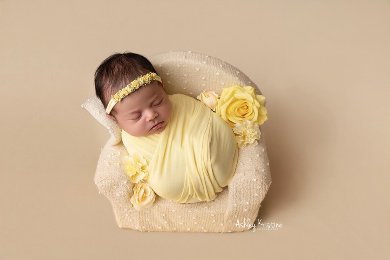 May include: A newborn baby wrapped in a soft yellow blanket, resting in a small, cream-colored chair. The baby wears a yellow floral headband. Yellow roses and other flowers decorate the chair. The background is a solid beige color.