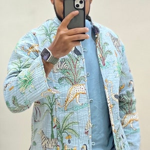 May include: A light blue quilted jacket with a jungle print featuring zebras, leopards, palm trees, and toucans. The jacket has a button closure and a light blue t-shirt underneath.