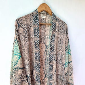 May include: A patterned kimono-style jacket in shades of beige, teal, and navy blue hangs on a wooden hanger. The fabric features an intricate paisley design. The jacket is open in the front, revealing the inner pattern.