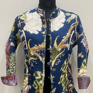 Może przedstawiać: Niebieska pikowana kurtka z kwiatowym nadrukiem. Kurtka ma kołnierz typu „mandarynka”, zapięcie na guziki i długie rękawy. Rękawy mają kontrastowe wykończenie z tkaniny.