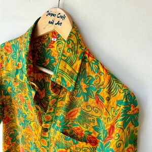 May include: A yellow, green and orange short-sleeved shirt with a floral pattern. The shirt has a button-up front and a pocket on the left side. The shirt is hanging on a wooden hanger with the text "Jaipur Craft and Art" printed on it.