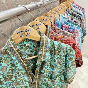 May include: A collection of teal blue and green floral print shirts hanging on wooden hangers with the text "Jaipur Craft and Art" printed on the hangers.