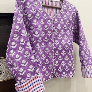 May include: A purple quilted jacket with a white floral print and a striped lining. The jacket has a button closure and long sleeves with striped cuffs.