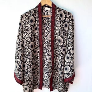 May include: A floral print kimono-style jacket. The jacket features a black and cream floral pattern with a red trim along the edges and down the front. The jacket is hanging on a wooden hanger against a white background.
