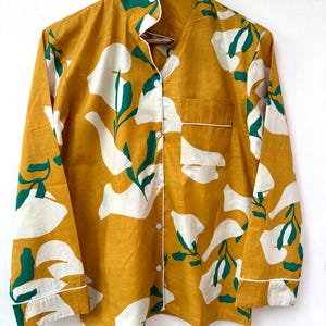 Puede incluir: Una camisa de manga larga con botones, fondo amarillo mostaza y estampado floral blanco y verde.