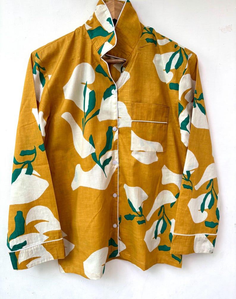 Puede incluir: Una camisa de manga larga con botones, fondo amarillo mostaza y estampado floral blanco y verde.
