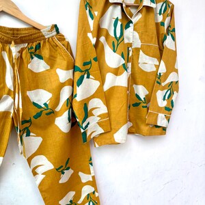 Puede incluir: Un conjunto de pijama amarillo mostaza y blanco con estampado floral. La parte superior tiene mangas largas y un cierre de botones en la parte delantera. Los pantalones tienen una cintura con cord&oacute;n.