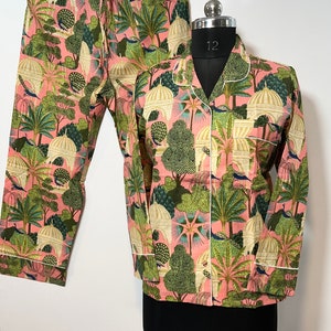 May include: Pink pajama set with a green and white print of peacocks, trees, and buildings. The top has a button-up front and long sleeves. The pants have an elastic waistband.