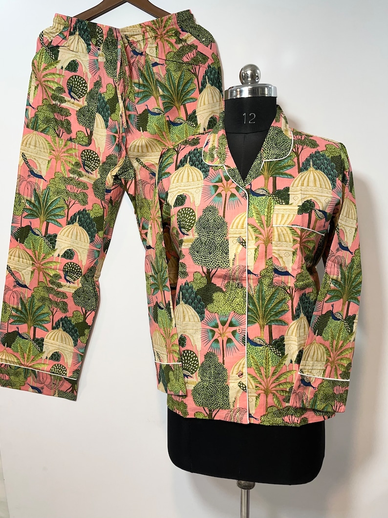 May include: Pink pajama set with a green and white print of peacocks, trees, and buildings. The top has a button-up front and long sleeves. The pants have an elastic waistband.