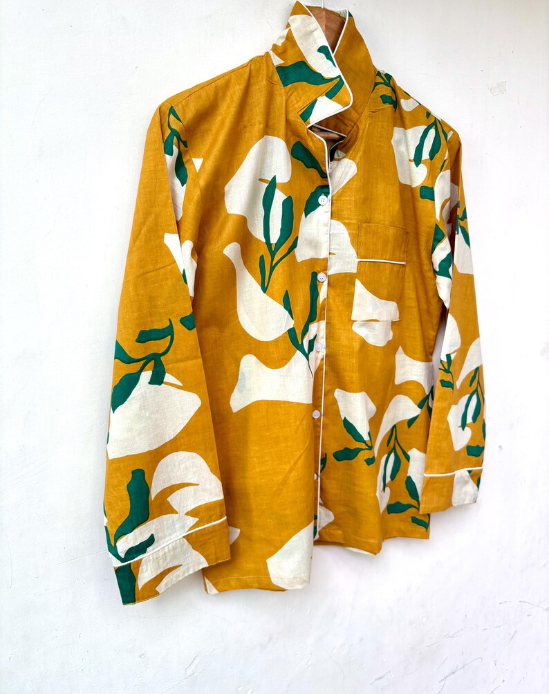 Puede incluir: Una camisa de manga larga de color amarillo mostaza con un estampado floral abstracto blanco y verde. La camisa tiene un cierre de botones en la parte delantera y un cuello.