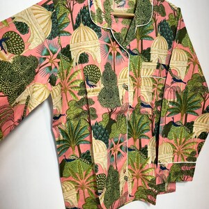 May include: A pink pajama top with a white trim and a print of peacocks, trees, and buildings. The label on the top reads "Acofab".