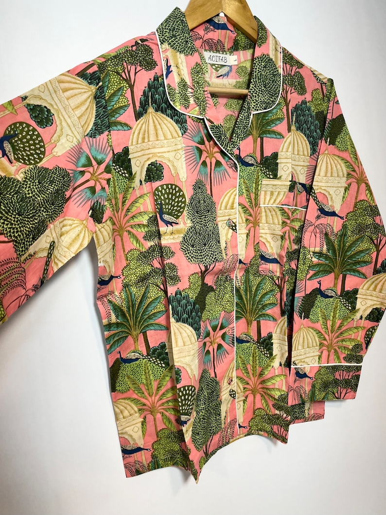 May include: A pink pajama top with a white trim and a print of peacocks, trees, and buildings. The label on the top reads "Acofab".