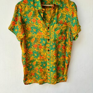 May include: A yellow, green and orange floral print short-sleeved button-up shirt with a pocket on the left side. The shirt has a collar and is hanging on a wooden hanger with the text "Jaipur Craft and Art" printed on it.