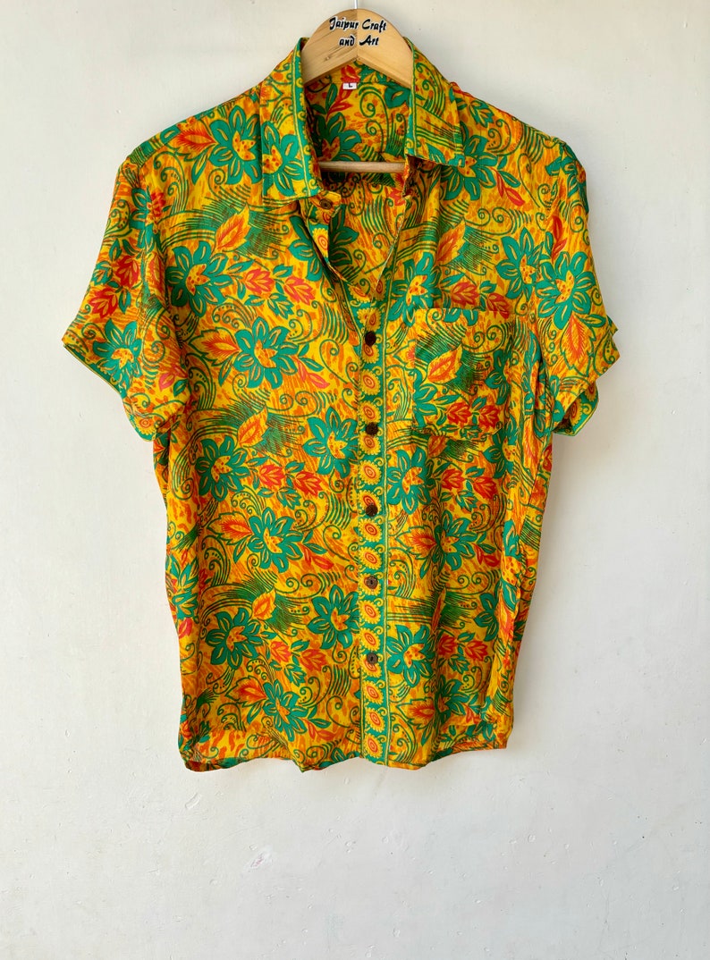 May include: A yellow, green and orange floral print short-sleeved button-up shirt with a pocket on the left side. The shirt has a collar and is hanging on a wooden hanger with the text "Jaipur Craft and Art" printed on it.