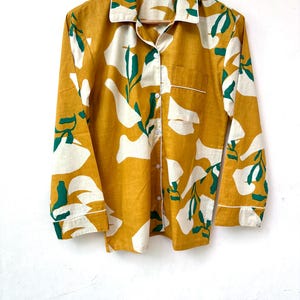 Puede incluir: Una camisa de manga larga de color amarillo mostaza con un estampado floral abstracto blanco y verde. La camisa tiene un cierre de botones en la parte delantera y un peque&ntilde;o bolsillo en el pecho izquierdo.