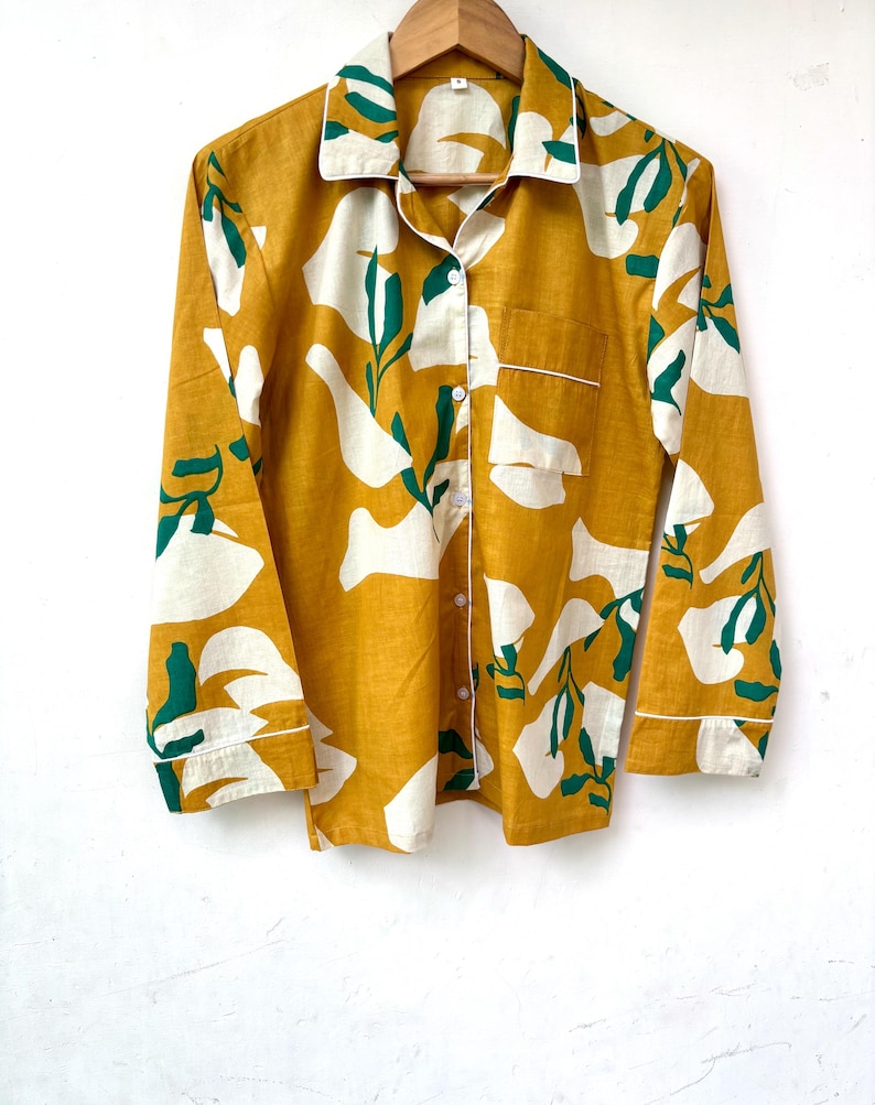 Puede incluir: Una camisa de manga larga de color amarillo mostaza con un estampado floral abstracto blanco y verde. La camisa tiene un cierre de botones en la parte delantera y un peque&ntilde;o bolsillo en el pecho izquierdo.