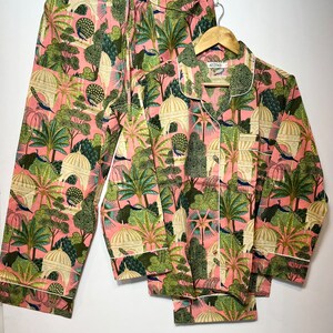 May include: A pink pajama set with a green and white print of peacocks, trees, and domes. The set includes a long-sleeved shirt with a button-up front and matching pants.