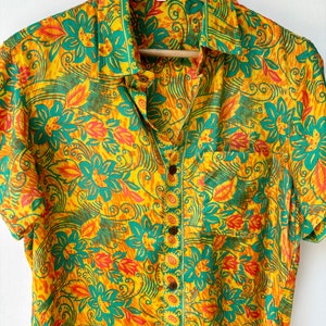 May include: A yellow, green and orange short-sleeved button-up shirt with a floral print. The shirt has a pocket on the left side and a button-up closure.