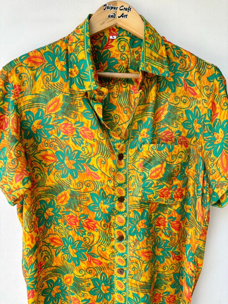 May include: A yellow, green and orange short-sleeved button-up shirt with a floral print. The shirt has a pocket on the left side and a button-up closure.