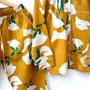 Puede incluir: Un conjunto de pijama amarillo mostaza con estampado floral abstracto blanco y verde. La parte superior tiene mangas largas y cierre de botones en la parte delantera. El pantal&oacute;n tiene una cintura el&aacute;stica.